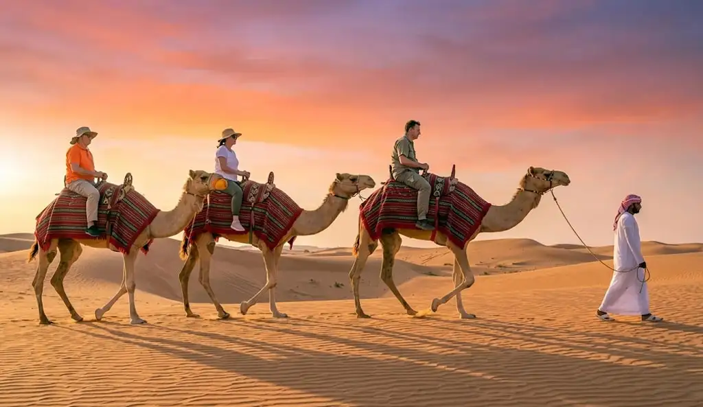 Tourists enjoying a sunset camel trekking tour in the Abu Dhabi desert, led by a local guide in traditional attire across sand dunes.