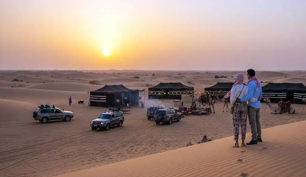 A panoramic view of an Evening Desert Safari Abu Dhabi Experience at sunset, showing a couple on a dune overlooking 4x4 vehicles, camels, and a traditional Bedouin camp as the sky turns orange.