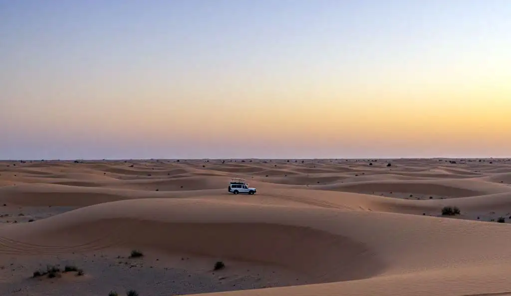 A breathtaking view of the vast, golden dunes of Al Khatim at sunset, showcasing the serene and remote environment of an Evening Desert Safari Abu Dhabi Experience with a single 4x4 vehicle in the distance.