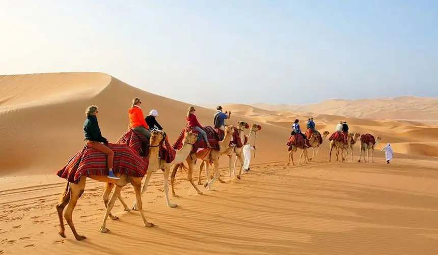 A group of tourists in bright clothing rides a line of camels over Abu Dhabi desert dunes.