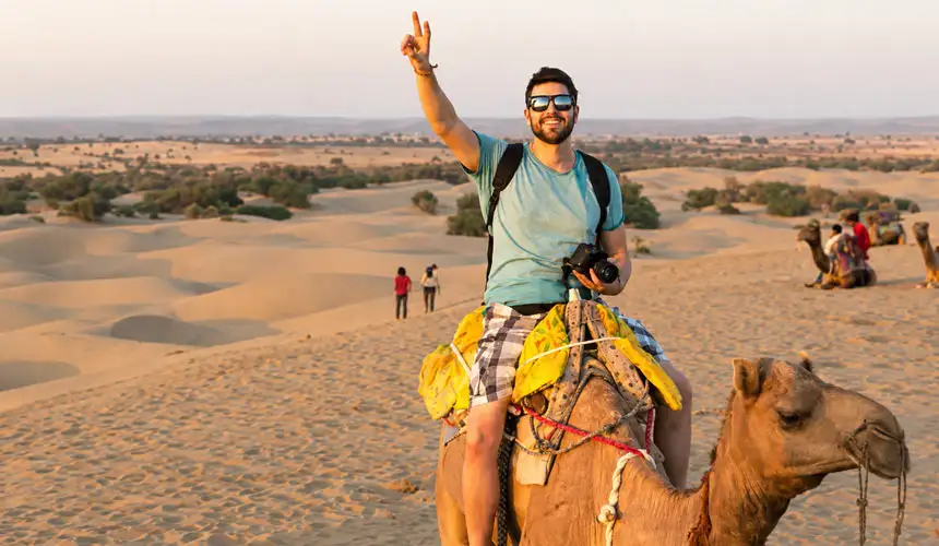 A smiling man in a teal shirt and sunglasses rides a camel, waving a peace sign against a Abu Dhabi desert backdrop.