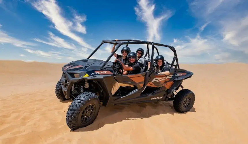 Happy group of four friends posing in a black and orange Polaris RZR 1000 dune buggy in the stunning Abu Dhabi desert under blue skies during their desert safari.