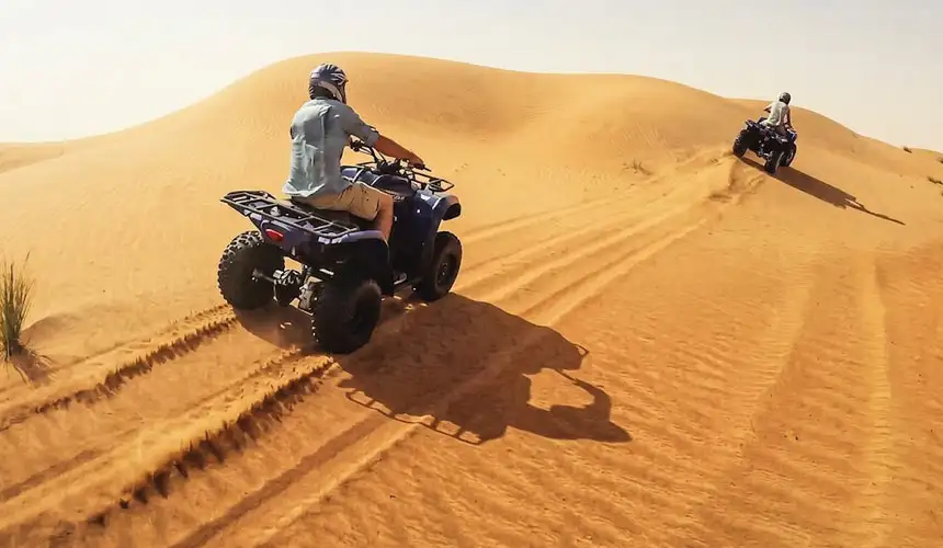 Exciting quad bike tour in Abu Dhabi desert with two riders on powerful red ATVs racing across golden sand dunes, kicking up sand under a bright blue sky. Perfect desert adventure experience in Abu Dhabi.