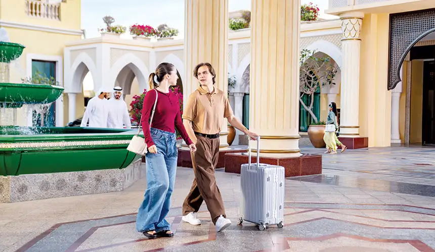 Young couple walking side by side with rolling suitcases through an elegant outdoor arcade with columns, fountains, and palm trees in a luxury Middle Eastern resort.