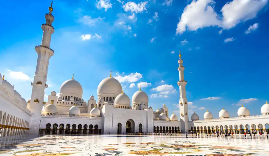 Grand view of the Sheikh Zayed Grand Mosque in Abu Dhabi under bright blue sky, featuring large white marble domes, tall minarets, and intricate floral courtyard design.
